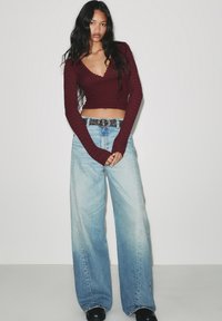 Burgundi hosszú ujjú, bordázott cropped top oversized világoskék, fakó farmernadrággal párosítva, fekete stúdás övvel, fekete cipővel kiegészítve.