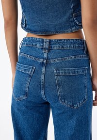 Jeans de mezclilla azul de cintura alta con un ajuste ligeramente holgado, que presentan bolsillos de parche y un acabado texturizado. La vista trasera muestra detalles de costura.