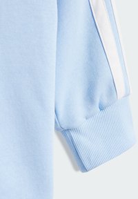 Light blue sweatshirt fabric with ribbed cuffs. White side stripes highlight the design. Smooth texture with a slightly stretchy fit.