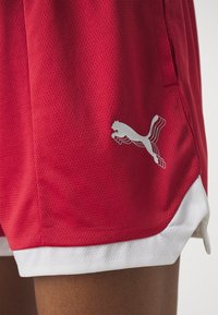 Pantalones cortos deportivos rojos hechos de tela de malla transpirable, con acentos blancos y un patrón texturizado. Logo aplicado en el lado inferior.