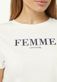 Witte katoenen t-shirt met korte mouwen, featuring vetgedrukte donkerblauwe tekst "FEMME," en "komma," gedrukt op de voorkant. Gladde textuur.