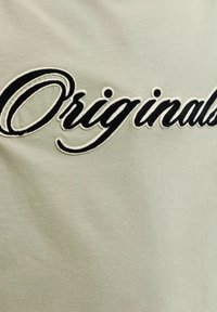 Μαύρο καλλιγραφικό κείμενο «Originals» κεντημένο σε φόντο από ανοιχτό πράσινο ύφασμα, που τονίζει το στυλ και τη σήμανση της μάρκας.