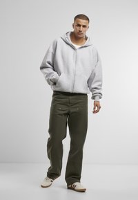 Graue Zip-Up-Hoodie mit lockerer Passform, kombiniert mit olivgrünen, weit geschnittenen Hosen, die sichtbare Taschen und Nähte aufweisen. Weiße Sneaker vervollständigen den Look.