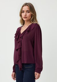 Blusa color bordeaux con scollo arricciato e maniche lunghe. Tessuto morbido e vestibilità leggermente ampia, abbinata a jeans scuri.