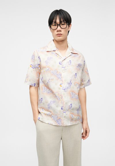 Jeune homme portant de grandes lunettes noires, une chemise à manches courtes claire à boutons avec un motif de papillons pastel, et un pantalon beige devant un fond uni.