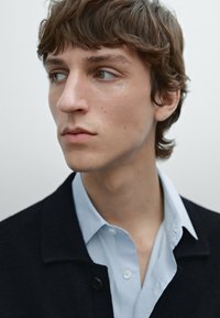 Black wool blazer over a light blue shirt, featuring a button and collar, with a close-up of a male model's neutral expression.