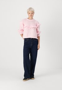Pull en laine rose avec des volants, manches longues et col rond, associé à un jeans ample bleu foncé et des chaussures noires.