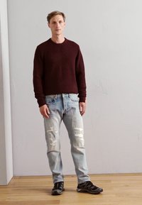 Pull en tricot bourgogne, jean délavé bleu clair avec des patches visibles, et chaussures de sport noires, sur un fond neutre.