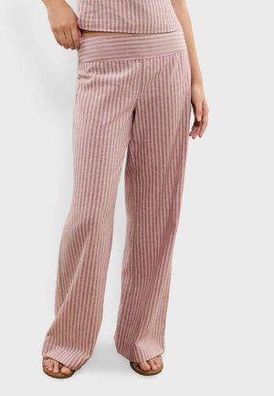 Femme portant un pantalon large à rayures roses et marron avec un haut assorti et des sandales marron, debout devant un fond uni.
