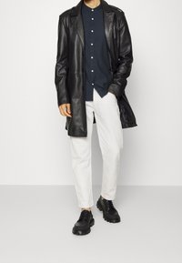 Cappotto in pelle nera sopra una camicia button-up blu, pantaloni bianchi e scarpe nere chunky. Il cappotto ha una texture liscia e una vestibilità lunga.