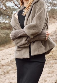 Cardigan marrón claro y suave al tacto, con mangas anchas y cierre de un solo botón, usado sobre un vestido negro ajustado.