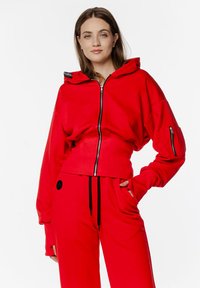 Czerwony krótki hoodie z zamkiem z przodu, z oversize'owymi rękawami i czarnymi detalami. Noszony z dopasowanymi czerwonymi dresami. Gładka tekstura.