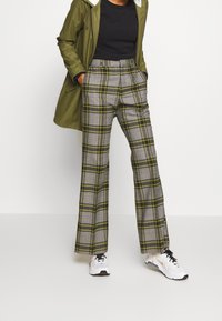 Pantalon gris à carreaux avec des accents jaunes, jambes larges, taille haute, porté avec un haut noir et une veste vert olive, assorti à des baskets blanches.