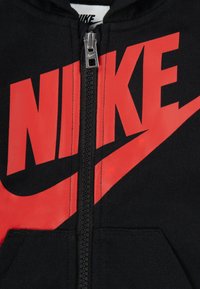 Crna zip-up hoodie s velikim crvenim Nike logotipom i dizajnom swoosh. Izrađena od mekanog materijala s teksturiranom površinom i prednjim džepovima.