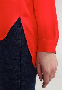 Camicia rossa con una texture liscia e setosa; presenta un orlo arrotondato e polsini con bottoni. Indossata con jeans in denim scuro.
