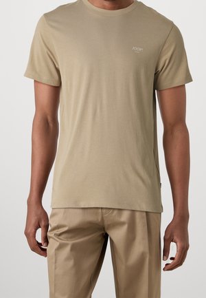 Homme portant un t-shirt beige à manches courtes avec petit logo "JOOP!" sur la poitrine et pantalon beige assorti sur fond uni.