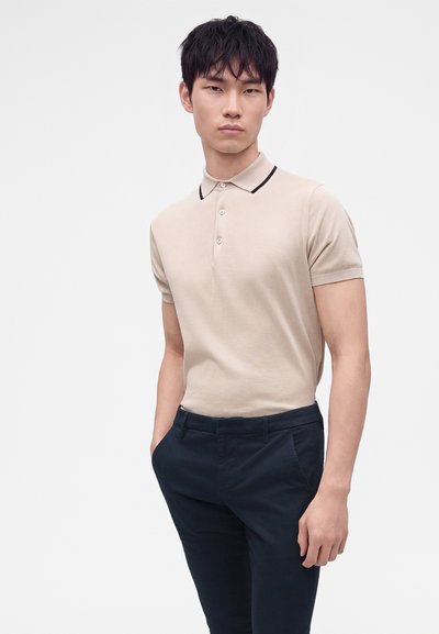 Homme portant un polo beige à manches courtes et un pantalon bleu marine foncé, debout les mains dans les poches devant un fond uni.