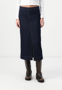 Denim suknja tamnoplave boje s prednjim prorezom, obrubljenim rubovima i kontrastnim žutim šavovima. Nosena s smeškim čizmama do gležnja.