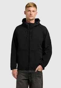 Jeune homme portant une veste zippée à capuche noire avec des poignets élastiques et un jean gris foncé, debout devant un fond clair uni.