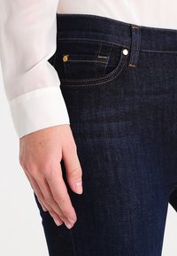 Tmavě modré džínové kalhoty s hladkým povrchem, přičemž mají kontrastní švy, malou přední kapsu a zlaté kovové detaily.