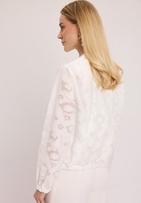 Biała, fakturowana bluzka z miękkim, falistym wzorem, elastycznym pasem i długimi rękawami. Lekka, przezroczysta tkanina zwiększa przewiewność.