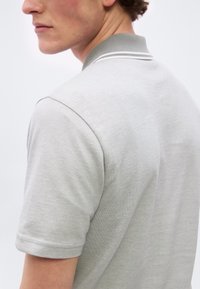 Homme portant un polo à manches courtes gris clair avec un col côtelé orné de deux rayures blanches, vu de dos et de profil.