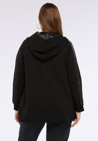 Donna con lunghi capelli castani che indossa un felpa nera con righe in satin sulle maniche, in piedi con le spalle rivolte verso uno sfondo bianco puro.