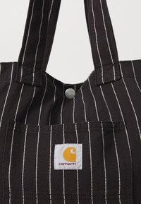Tote de lona negra con rayas verticales blancas, que cuenta con un cierre de broche metálico y un parche con el logo de Carhartt. Mangos dobles resistentes para llevar.