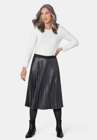Zwarte plissé midi-rok met glanzende textuur, brede tailleband, gecombineerd met een wit shirt met lange mouwen en zwarte enkelboots.