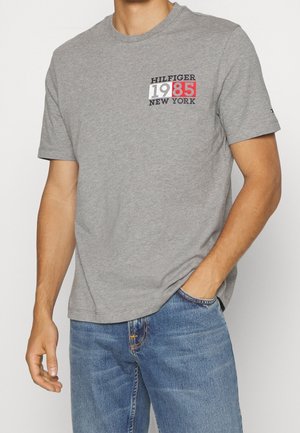 Mężczyzna ubrany w szarą koszulkę z krótkim rękawem z napisem "HILFIGER 1985 NEW YORK" na klatce piersiowej, w połączeniu z niebieskimi dżinsami.