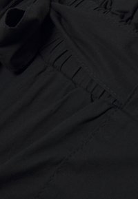Svart tyg med samlat elastiskt detalj, jämn textur och kontrasterande sömnad. Ytan visar en subtil lyster på vissa områden.
