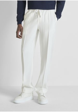 Uomo che indossa pantaloni bianchi pieghettati con coulisse e sneakers bianche, mani in tasca, con maglia a maniche lunghe blu navy visibile dalla vita in giù.