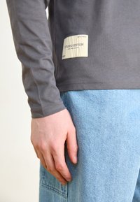 Gray long-sleeve shirt made of soft fabric featuring a stitched cream label with "STUDIO EDITION," paired with light blue denim.