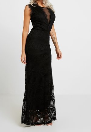 Kvinde i en ærmeløs, gulvlængde sort blondekjole med et gennemsigtigt V-udskæringspanel, stående mod en ensartet lys baggrund.