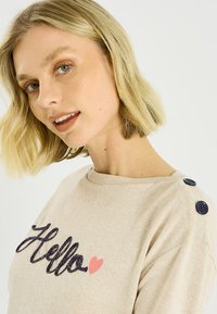 Suéter beige con estampado de "Hola" en azul oscuro y un corazón rosa. Diseñado con cuello redondo y detalles de botones azul marino en el hombro.