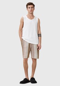 Weißes geripptes Tanktop kombiniert mit beigen und cremefarbenen gestreiften Shorts, die einen Kordelzug und eine strukturierte Oberfläche aufweisen. Schwarze Slipper sind sichtbar.