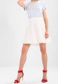 Bílá áčková sukně s vyšívaným designem, zkombinovaná s světle modrým tričkem s krátkým rukávem. Červené sandály na podpatku s chlupatými řemínky kolem kotníků.