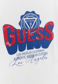 Biała koszulka z dużą czerwoną grafiką "GUESS", niebieskimi akcentami oraz okrągłym logo z napisem "EST. 1981" poniżej. Tekst: "Los Angeles California Autentyczna Amerykańska Odzież."