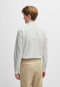 Helles, langärmliges Hemd mit kleinen Musterdetails, geknöpftem Kragen und weicher Textur. Kombiniert mit hellbeigen Hosen.