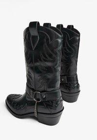 Črne usnjene kavbojke z zapletenim šivom, s konicami prstov in nizkim peto. Imajo okrasen pašček in podrobnosti z verigo.