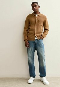 Uomo che indossa un cardigan beige con bottoni sopra una camicia bianca, jeans blu e sneakers bianche, in piedi contro un muro chiaro e semplice.