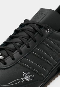 Black Adidas leather sneaker with black laces, three stitched stripes, and white floral embroidery near the toe area.