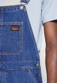 Modré džínové lacláče s nastavitelnými ramenními popruhy, kovovými doplňky a přední kapsou s nášivkou s logem Levi's.