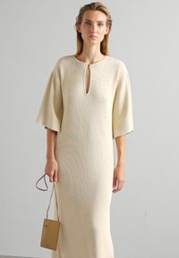 Μακρύ, μπεζ πλεκτό φόρεμα με κοντά, φαρδιά μανίκια. Περιλαμβάνει λαιμόκοψη με άνοιγμα και ομαλή υφή. Το μοντέλο κρατά μια μικρή, ταιριαστή τσάντα χεριού.