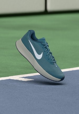 Μπλε παπούτσι για τρέξιμο Nike με λευκό σήμα Swoosh, που αιωρείται πάνω από μια πράσινη και μπλε αθλητική επιφάνεια κοντά σε μια λευκή γραμμή.