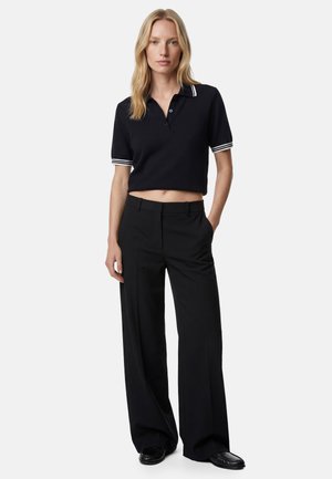 Kvinde står iført en sort cropped polo skjorte med hvid kant og sorte bukser med vide ben og sorte loafers.