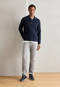 Tengerészkék, hosszú ujjú galléros póló, fehér gallér kiemelésekkel, világosszürke farmerrel és sötét színű sneakers cipőkkel párosítva. Az anyag simának tűnik, és laza szabású.