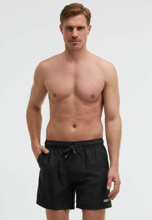 Sportlicher Mann steht mit einer Hand in der Tasche, trägt schwarze Badeshorts mit Kordelzug vor einem einfarbigen hellen Hintergrund.