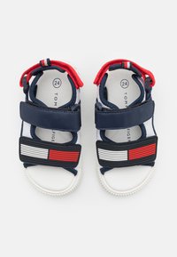 Marinblå och vita sandaler med röda detaljer, justerbara remmar och en texturerad sula. Har horisontella ränder i röd och vit på framsidan.