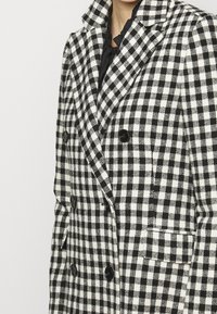 Dubbelgeknoopte jas in zwart-wit geruit patroon. Gemaakt van gestructureerde stof met reverskraag en twee voorzakken. Zwarte knopen.